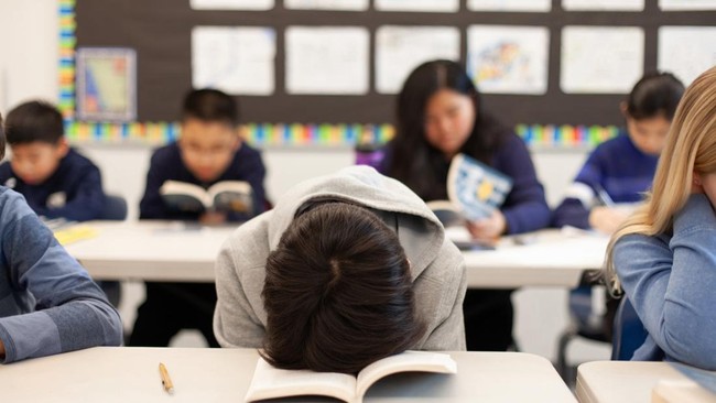 Elementary school junior high boy student sitting at desk in classroom with head in book in frustration sleeping tired exhaustion boredom