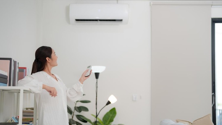 Young woman is using a remote control to adjust the air conditioner in a bright, modern living room with minimalist decor