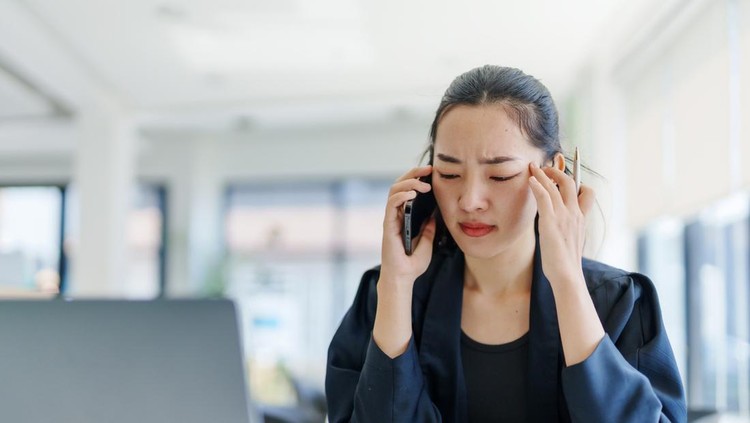 Young Asian businesswoman having a headache and massaging her temples while making a phone call in the office