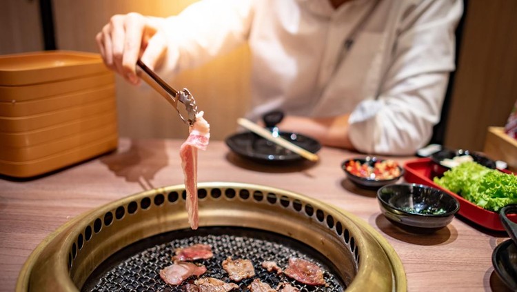Asian man using kitchen tongs grilling beef and bacon on grill plate. Eating Korean Barbecue buffet in the restaurant.