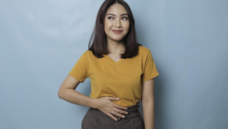 Pleased cheerful Asian woman keeps hand on belly feels full after delicious dinner dressed casually stands thoughtful against blue background.