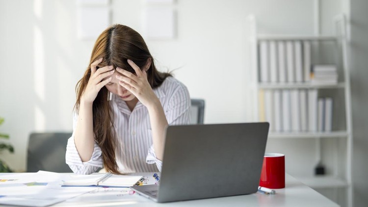 A woman is sitting at a desk with a laptop and a red cup. She is looking at the laptop with a worried expression on her face