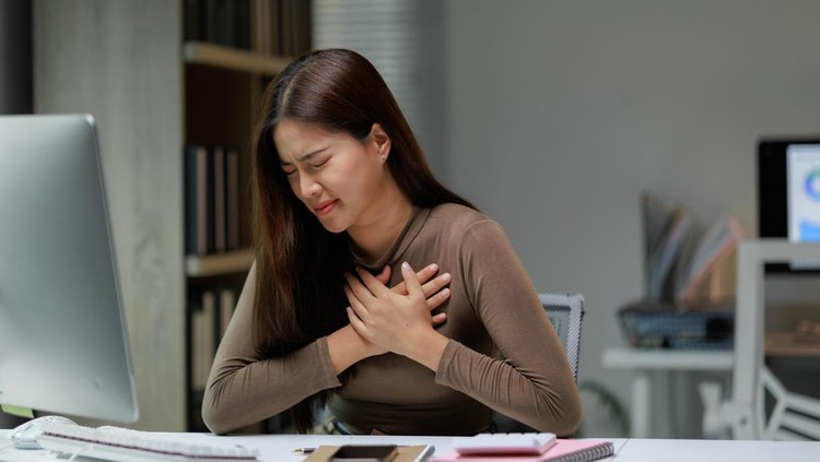 Young asian businesswoman clutching her chest in pain while working at her desk in a modern office
