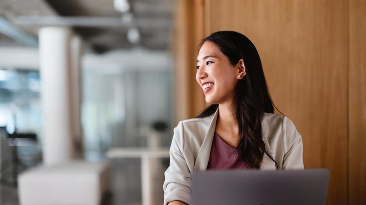 Ilustras pegawai atau karyawan di kantor Confident young Asian businesswoman smiling while sitting at a desk in a modern office setting, exuding positivity and success.