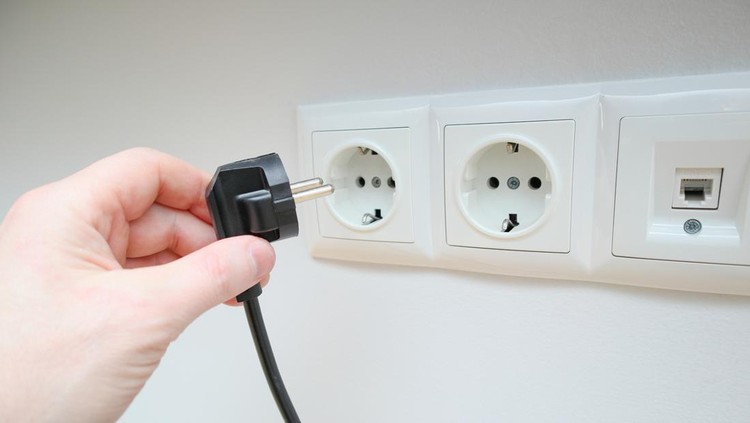 A man's hand holds an electrical plug next to the sockets on the white wall. Human hand inserts a plug into an electrical outlets. Hand puts plug in the socket