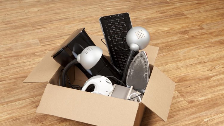 Cardboard box filled with an assortment of household equipment or appliances, ready for moving or a garage sale. Items in the box include a coffee maker,electric mixer, iron, small heater,computer keyboard, and lamps.