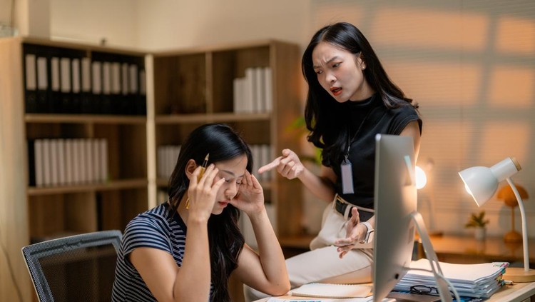 Young stressed asian businesswoman pressing palms against ears, blocking manager's criticism during workplace confrontation near office workstation