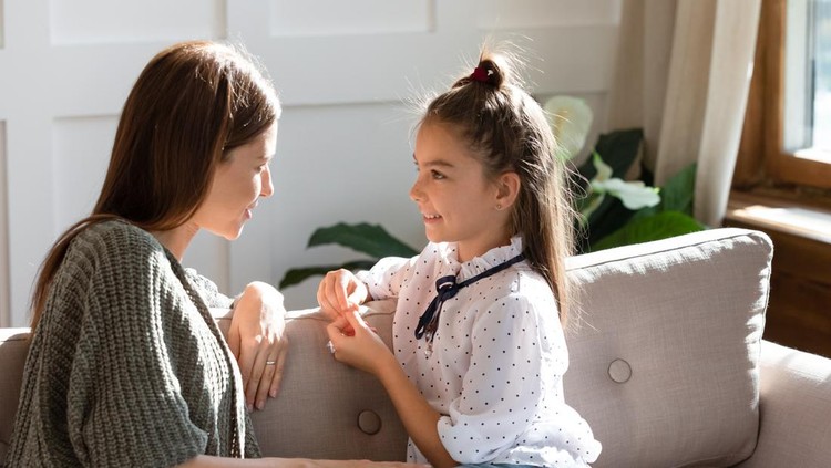 Young Caucasian mom sit on couch with small daughter talk share secrets gossip at home. Happy little girl child speak with mother or nanny enjoy tender intimate moment together. Bonding concept.