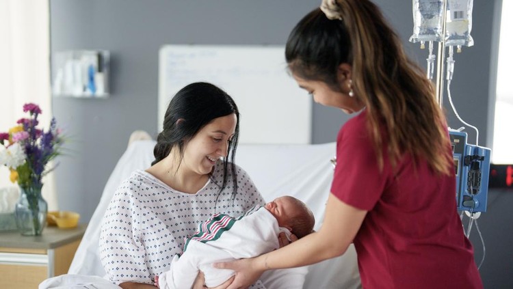 A female nurse of Asian decent hands over a newborn baby to his mother who is sitting up in her hospital bed shortly after giving birth.  The baby is swaddled tightly and appears content as the Mother embraces her baby.
