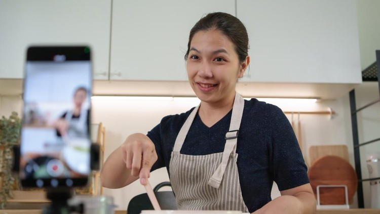 Cheerful Asian cook mixes food and smiles while creating recipe video content with smartphone for social media followers.
