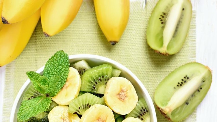 Kiwi and banana salad in bowl and fruits on white wooden table, top view