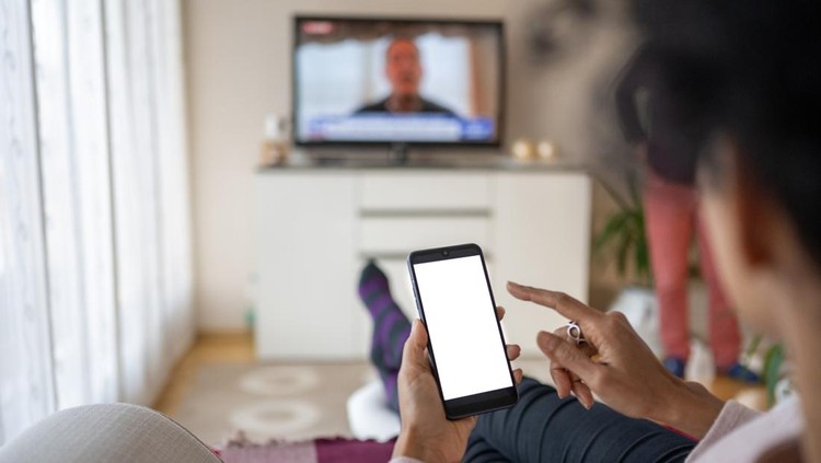 Over the shoulder view of a hispanic mixed race woman using a smart phone with a white screen while sitting on a sofa in the living room and watching TV&period;