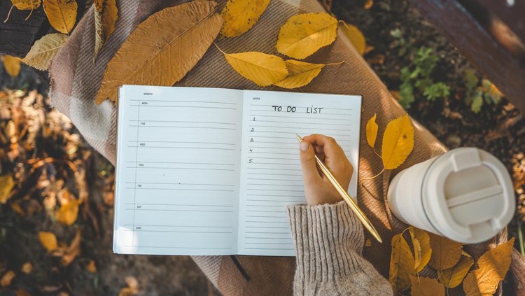 Hand writing in a planner with a gold pen&comma; surrounded by yellow autumn leaves&comma; a reusable cup&comma; and cozy textures&comma; creating a warm seasonal to-do list scene&period;