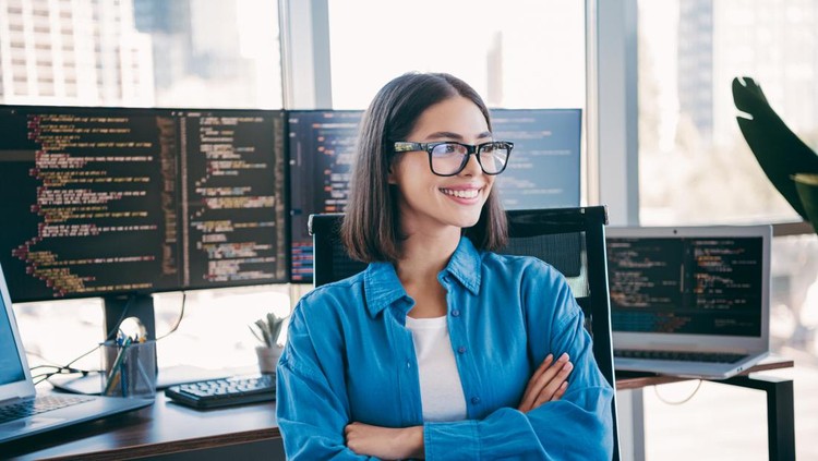 A cheerful female programmer sits at a busy desk surrounded by multiple computer monitors displaying code in a bright modern office setting&period; Her confident smile conveys professionalism and focus in a tech driven environment&period;