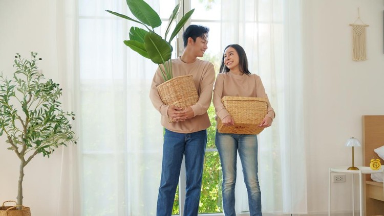 Moving house concept&comma; Asian couple smiling while holding woven baskets&comma; standing in a bright room with large windows and indoor plants&comma; preparing to organize their home together&period;