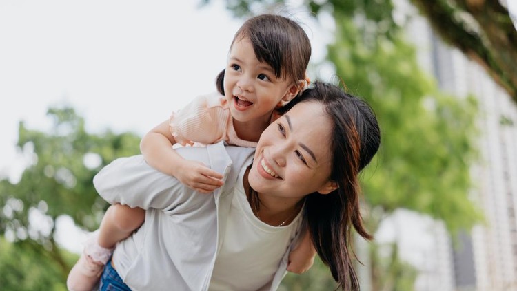 An Asian Chinese mother playing piggyback riding with her young daughter in a public park