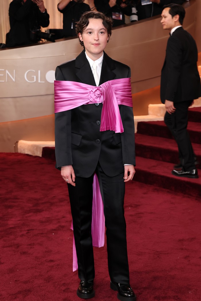 Sama-sama mengenakan tuxedo rancangan Prada, Bella Ramsey menampilkan pesona androgini segar dan unik. Setelan jas dan celana panjang dihiasi aksen&nbsp;shawl satin&nbsp;besar fuchsia yang disematkan seolah pita yang membungkus tubuhnya/ Foto: Courtesy of Prada/Getty Images/Amy Sussman