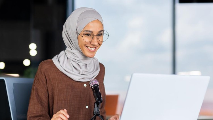 A young Muslim woman in a hijab is sitting in an office in front of a laptop and a microphone&period; records a podcast&comma; a blog&comma; talks on a video call&comma; gives an interview&comma; goes through an online interview&period;