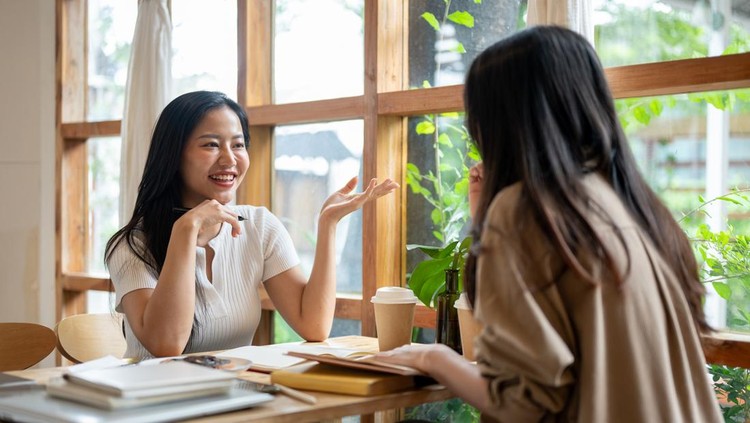 Two cheerful young Asian college students are enjoying talking and reading together in a minimalist coffee shop&period; people&comma; lifestyles&comma; university life&comma; leisure
