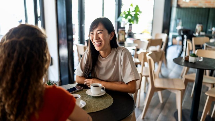 Two young women sharing laughter and engaging in a lively conversation while sipping coffee together at a bright&comma; modern cafe