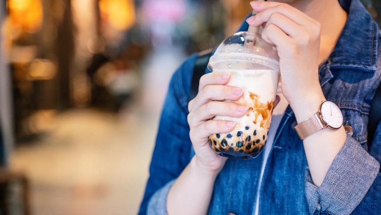 Young girl in denim jacket is drinking brown sugar flavored tapioca pearl bubble milk tea with glass straw in night market of Taiwan&comma; close up&comma; bokeh
