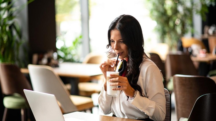 Young businesswoman in a white shirt drinking latte while looking at her laptop screen in a modern cafe&period;