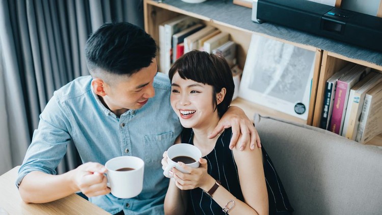 Loving young Asian couple chatting and having coffee while relaxing in living room at home