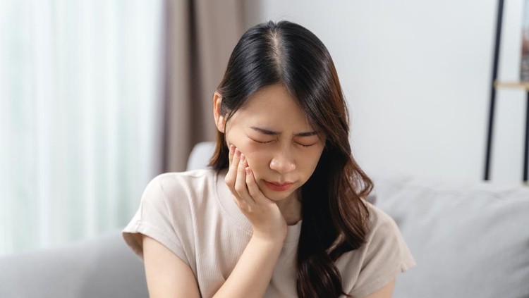 The woman with a toothache is holding her cheek in pain while sitting on the sofa in the living room at home&period; Healthcare and medical concept&period;