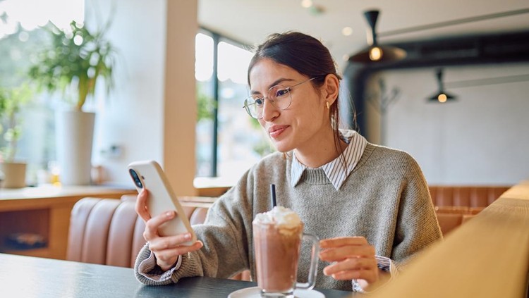 Young woman relaxing at a cafe&comma; holding a smartphone&comma; interacting with digital content while sipping a hot chocolate