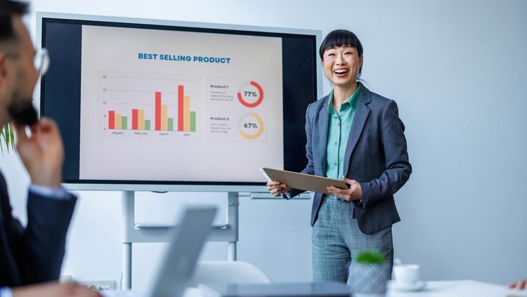 Businesswoman stands in a meeting room presenting sales data on a chart&comma; colleagues listening