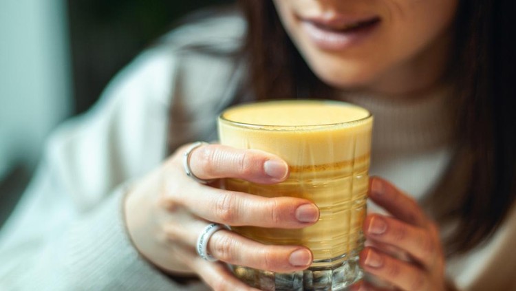 Young caucasian woman in warm sweater is smiling and holding a glass of hot turmeric milk on table of coffee shop&period; Enjoyment of drinking hot beverage near window on weekend&period; Copy space