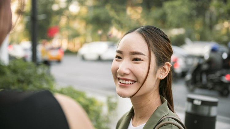 Young asian woman smiling and looking at her friend while having a conversation on a city street&comma; with blurred cars and trees in the background