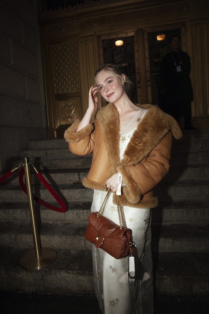 Di tengah dinginnya kota New York, Elle Fanning membuktikan gaya musim dingin nggak harus monoton. Fur jacket yang menumpuk&nbsp;gaun panjang dengan sheer overlay bermotif bintang&nbsp;menegaskan kesan feminin dan glamor, tapi punya playful twist. Terlebih dengan tas Tabby dengan gantungan miniatur buku dalam genggaman/ Foto: Dok. Coach