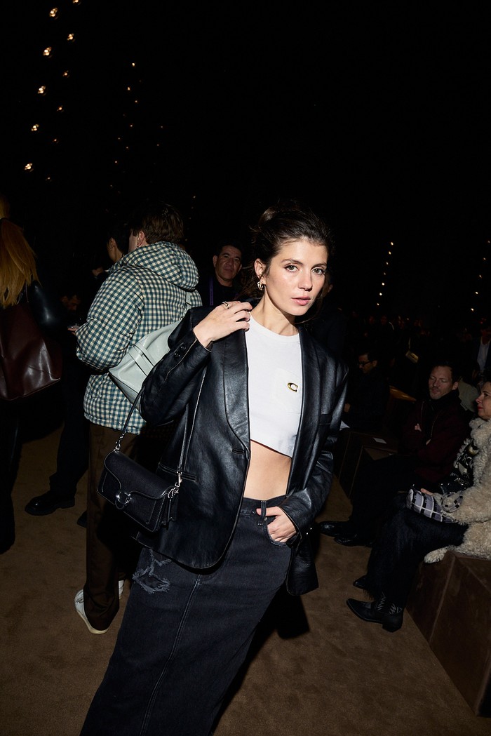 Emily Bader menampilkan gaya black & white. Cropped t-shirt putih melapisi blazer kulit hitam yang senada dengan rok span. Tas Tabby hitam melengkapi look-nya./ Foto: Dok. Coach