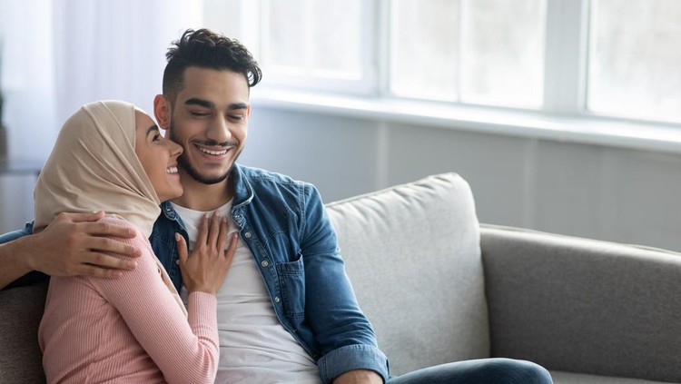 Happy middle-eastern family husband and wife cuddling on couch at home&comma; embracing&comma; talking and laughing&comma; panorama with copy space&period; Cheerful arab couple enjoying time together