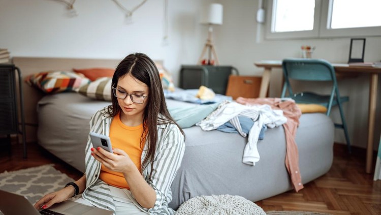 Young beautiful woman using phone and laptop in her messy bedroom