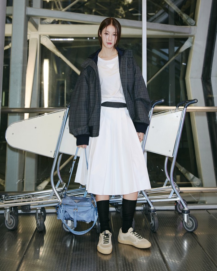 Namtan Tipnaree Weerawatnodom terlihat lebih edgy dalam outfit bersiluet oversized yang longgar. Perpaduan antara kaus dan midi skirt putih dengan jaket abu-abu bermotif tartan seluruhnya dalam rona netral membuat tas birunya standout/ Foto: Dok. Prada