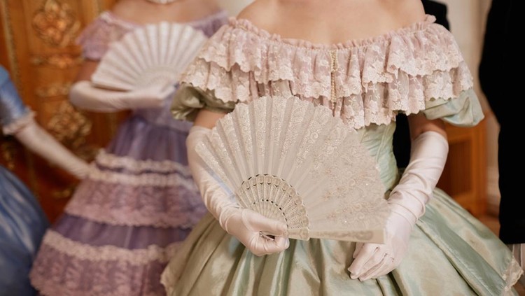 Closeup of ladies wearing big ballgowns holding fans while waiting for partners during ball in palace&comma; copy space