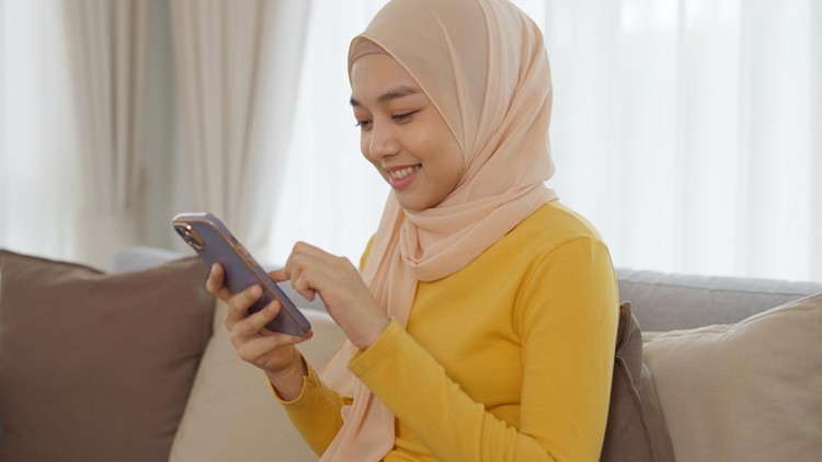 Young beautiful Muslim woman wearing hijab headscarf is using smartphone and selfie sitting on sofa at home&period;