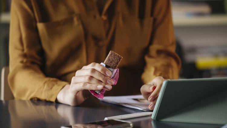 An anonymous businesswoman sitting at the office&comma; eating a protein bar while video calling with her colleagues