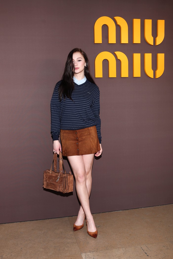 Lola Tung tampil preppy dengan menumpuk kemeja biru dan V-neck sweater motif garis. Ia mengombinasinya dengan mini skirt suede yang senada dengan pointy p/ Foto: Dok. Miu Miu