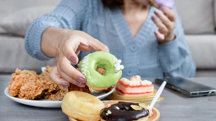 Woman reaching for a donut from a table full of junk food&comma; showing signs of sugar cravings&comma; overeating&comma; and unhealthy eating habits
