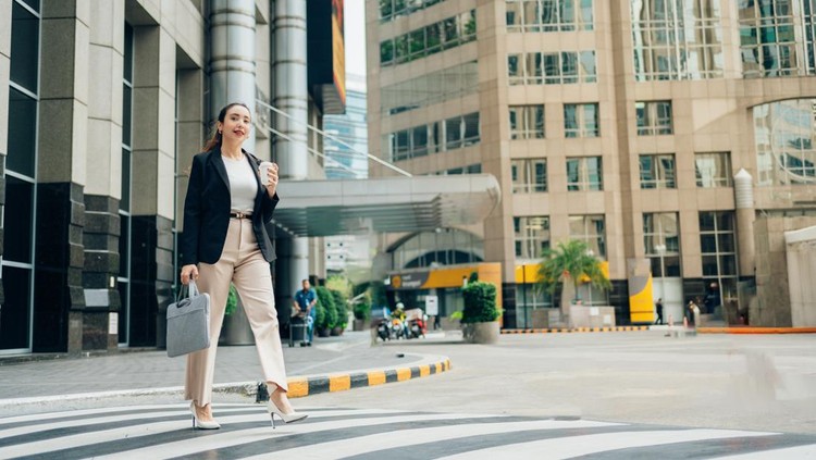 Young businesswoman walking across a pedestrian crossing in the financial district&comma; holding a takeaway coffee and a laptop bag&comma; going to work
