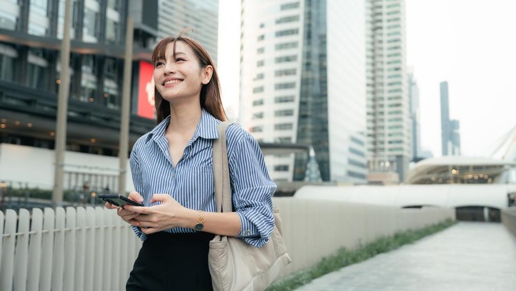Happy young businesswoman walking to work&comma; texting on her smartphone as she navigates the morning rush&comma; confirming meeting agendas&comma; reading real-time updates&comma; and syncing with her remote team&comma; balancing efficiency with ease&comma; embodying the rhythm of modern work-life balance