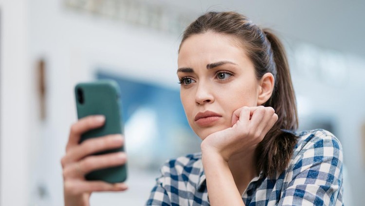 Close up of a depressed Caucasian young woman sitting at home&comma; reading some bad news on her smart phone&comma; feeling sad and worried