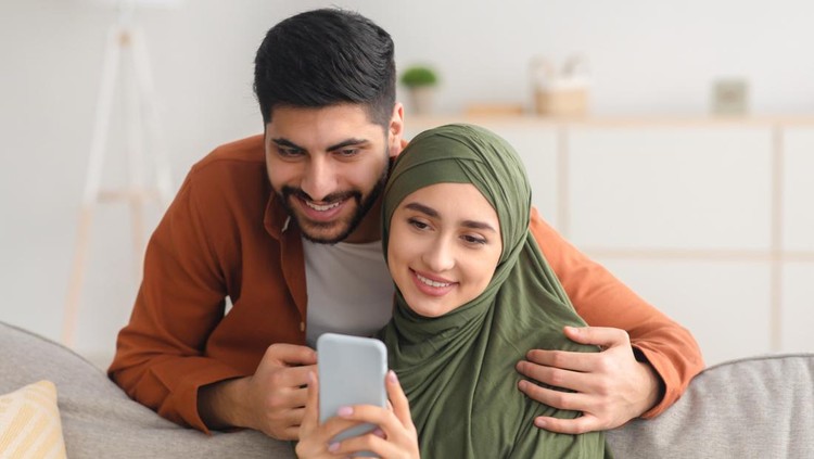 Happy Middle Eastern Spouses Using Mobile Phone Texting And Websurfing Sitting On Couch At Home&period; Modern Communication And Technology&comma; Great Application For Cellphone Concept