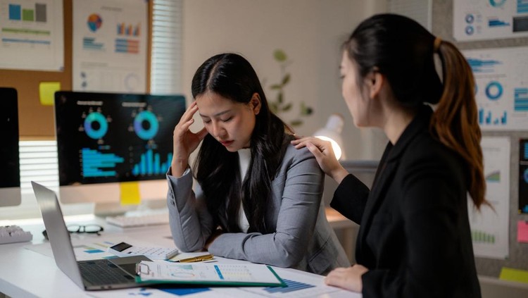 Professional Asian businesswomen supporting stressed colleague working late on laptop in dimly lit corporate workspace
