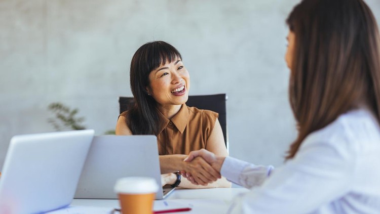 Professional interaction between two women shaking hands in an office setting&comma; highlighting business collaboration and partnership&period; Laptops and coffee cups are visible in the workspace&period;