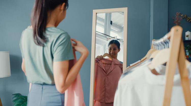 Beautiful attractive Asia lady choosing clothes on clothes rack dressing looking herself in mirror in living room at house&period; Girl think what to wear casual shirt&period; Lifestyle women relax at home concept&period;