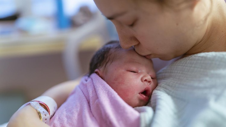 Shortly after birth a newborn baby boy is wrapped tightly in a swaddling blanket and laid on the bed to rest&period;  The Asian baby has a full head of dark hair and has his eyes closed as he sleeps peacefully&period;  His Mother is leaning in closely over him to watch as he sleeps&period;